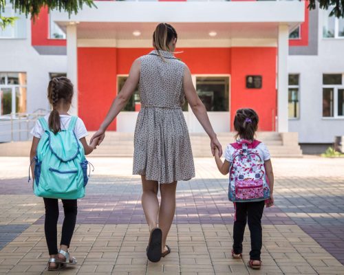 Back to school education concept with girl kids, elementary students, carrying backpacks going to class on school first day holding hand in hand together walking up building stair happily
